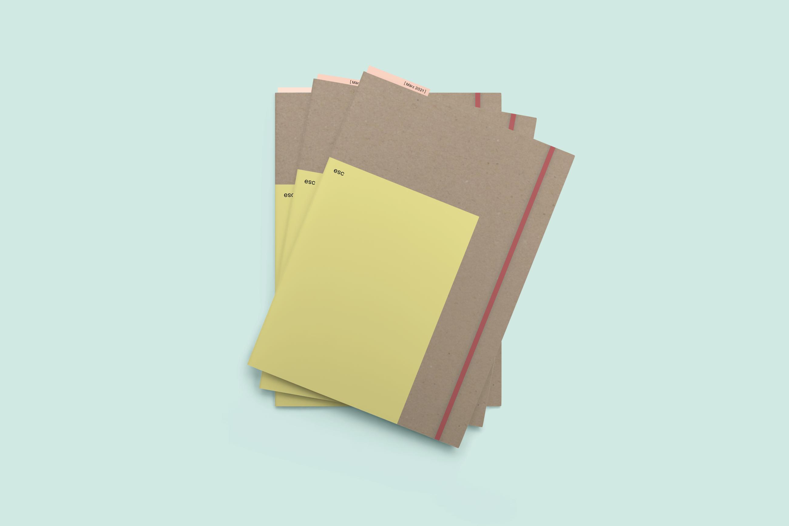 Mockup Datei von 3 dünnen Büchern mit einem Graupappe Umschlag, einen kleineren gelben Umschlag mit der Aufschrift "esc" und einem roten Gummiband
